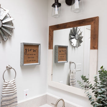 A bathroom with a farmhouse-style design features a wooden-framed mirror, a framed quote on the wall, a striped hand towel, and decorative metal windmill wall art. The mirror reflects the framed quote and windmill decor, enhancing the rustic aesthetic.