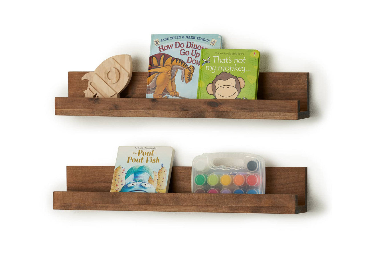 Two brown wooden floating shelves are mounted on a white wall, displaying children's books and a wooden rocket. The top shelf holds two picture books and a wooden rocket, while the bottom shelf holds a picture book and a watercolor paint set.
