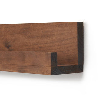 A close-up shows a long, narrow, dark brown wooden floating shelf mounted on a white wall. The shelf has a raised front lip to hold items in place.