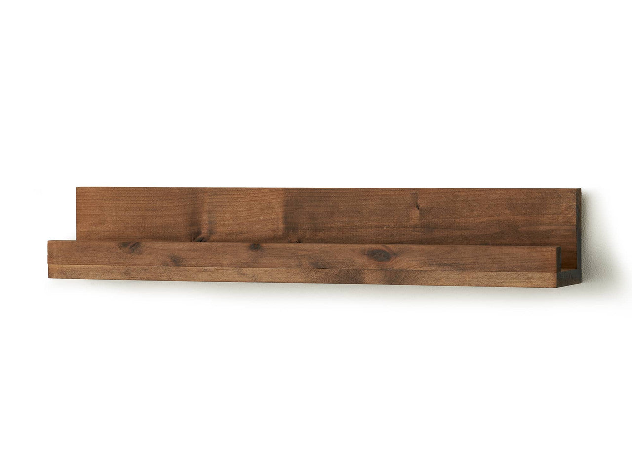 A horizontal wooden floating shelf with a warm brown stain is mounted on a white wall. The shelf has a raised back and a small lip at the front, designed for displaying books or other small items.