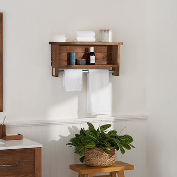 A bathroom shelf made of dark wood is mounted on a white wall above two hanging white towels. The shelf holds various bathroom items, including rolled white towels, a white container, a metallic can, a small blue cup, and a brown bottle.