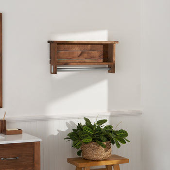 A bathroom features a wooden framed mirror to the left and a matching wooden shelf with a metal towel bar mounted on a white wall. Below the shelf, a potted plant sits on a small wooden stool near a window showing a bright outdoor scene.