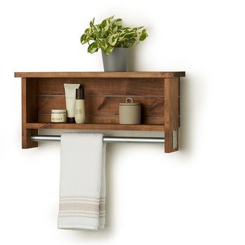 A wooden towel bar with shelves, identified is mounted on a white wall. The top shelf holds a small potted plant, while the lower shelf contains a jar and two bottles; a white towel hangs from the silver-toned bar below.