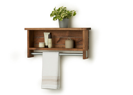 A wooden towel bar with shelves, identified is mounted on a white wall. The top shelf holds a small potted plant, while the lower shelf contains a jar and two bottles; a white towel hangs from the silver-toned bar below.
