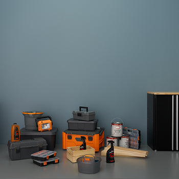 A still-life photo shows a collection of garage and workshop supplies, featuring a large orange and black shop vacuum, various storage bins, toolboxes, and cleaning products. To the right is a black, two-door storage cabinet with a light wood top, contrasting with the dark gray wall behind the items.