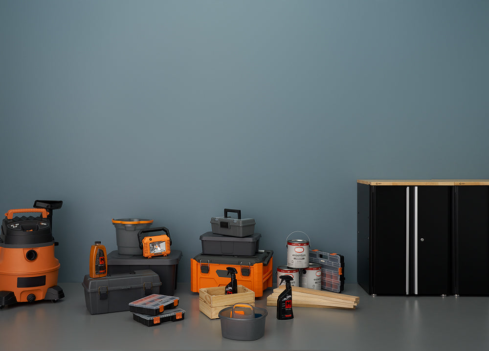 A still-life photo shows a collection of garage and workshop supplies, featuring a large orange and black shop vacuum, various storage bins, toolboxes, and cleaning products. To the right is a black, two-door storage cabinet with a light wood top, contrasting with the dark gray wall behind the items.