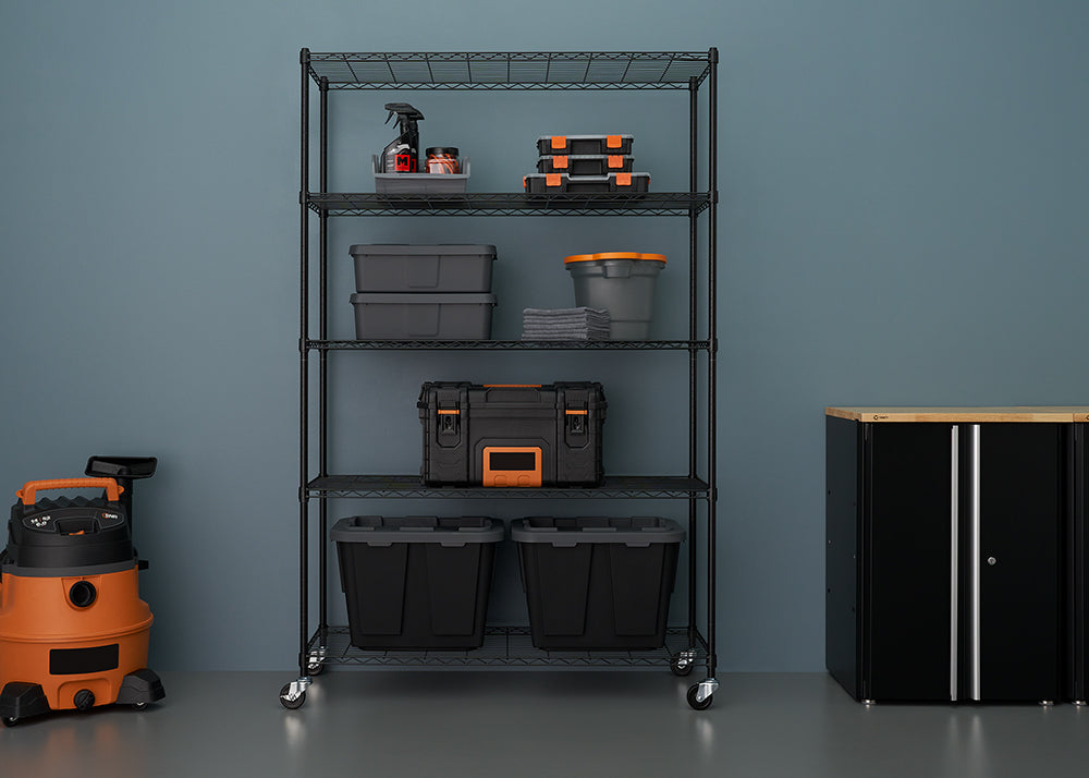 A tall, five-tier black wire shelving unit with casters is centered against a muted blue wall, fully organized with various black and grey storage bins, toolboxes, and cleaning supplies. To the left is an orange and black wet/dry vacuum, and to the right is a black cabinet with a light wood top.