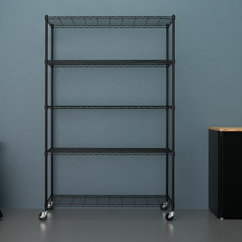 
A five-tier, black wire shelving unit stands centered against a blue-gray wall, equipped with casters for mobility. To its left is an orange and black wet/dry vacuum, and to the right is a black storage cabinet with a wooden top.