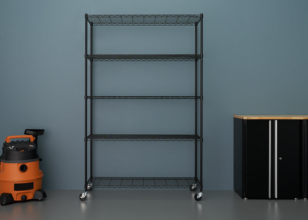 
A five-tier, black wire shelving unit stands centered against a blue-gray wall, equipped with casters for mobility. To its left is an orange and black wet/dry vacuum, and to the right is a black storage cabinet with a wooden top.