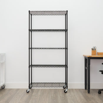 A tall, black, five-shelf wire shelving unit with wheels is centered against a white wall in an office setting. To the left is a white rolling cart with a coffee maker, and to the right is a wooden desk and a black office chair.