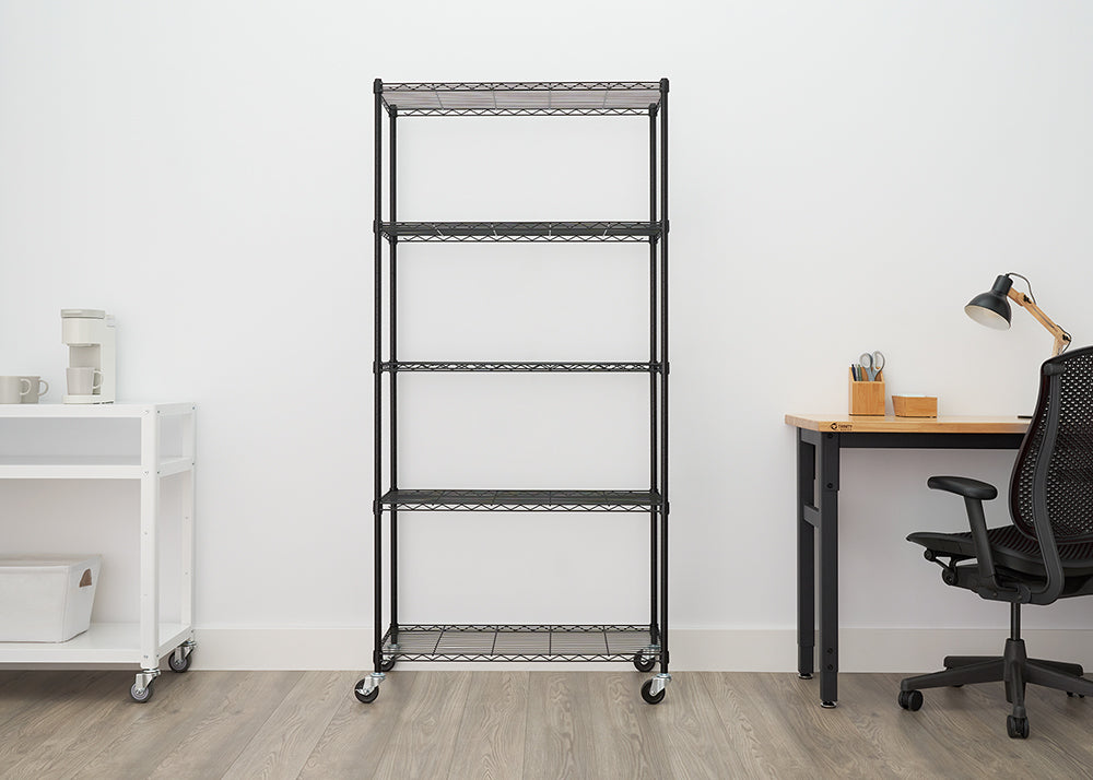 A tall, black, five-shelf wire shelving unit with wheels is centered against a white wall in an office setting. To the left is a white rolling cart with a coffee maker, and to the right is a wooden desk and a black office chair.