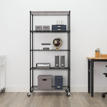 A five-tier, black wire shelving unit on wheels is centered in an office setting, stocked with various office supplies like folders, storage boxes, a globe, and a small printer. To the left is a smaller white rolling cart with a coffee maker, and to the right is a black desk with an ergonomic chair and a wooden-base task lamp.