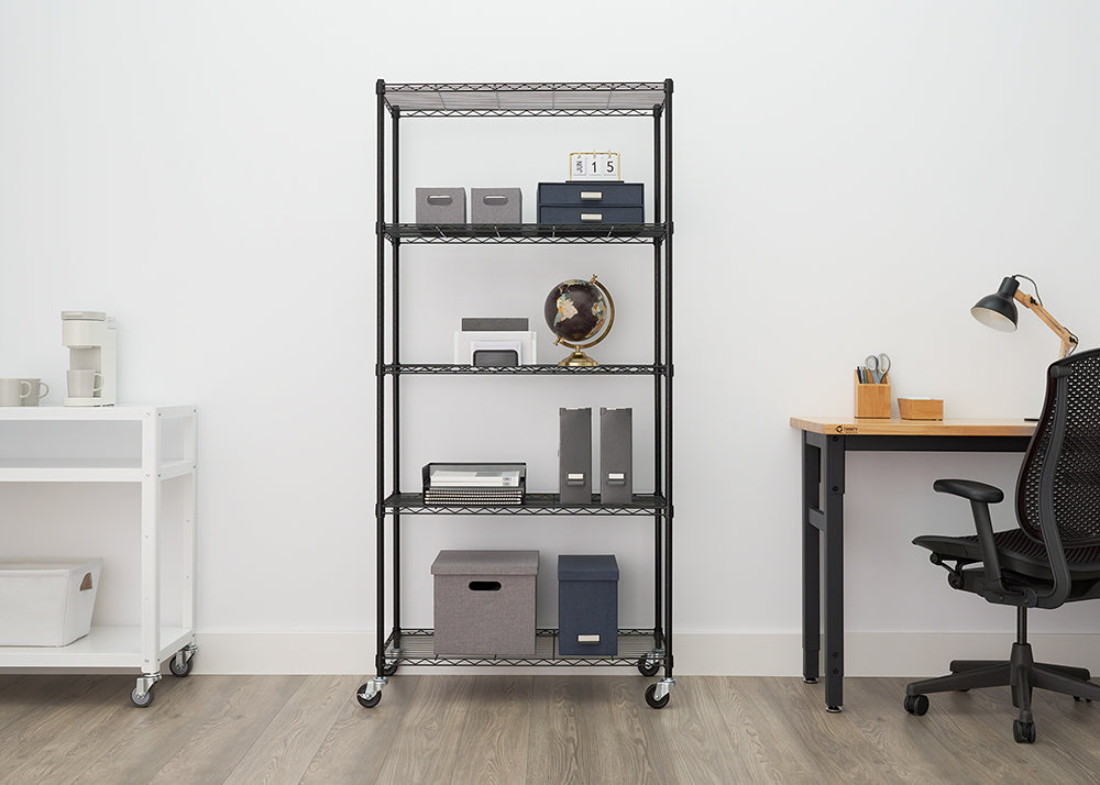 A five-tier, black wire shelving unit on wheels is centered in an office setting, stocked with various office supplies like folders, storage boxes, a globe, and a small printer. To the left is a smaller white rolling cart with a coffee maker, and to the right is a black desk with an ergonomic chair and a wooden-base task lamp.