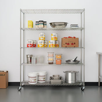 A five-shelf chrome wire shelving unit on casters is centered against a white wall and stocked with various food storage and cooking supplies, including canned goods, dry goods, mixing bowls, and pots. To the left are two cardboard boxes, and a stainless steel utility table is visible on the right next to a wall clock.