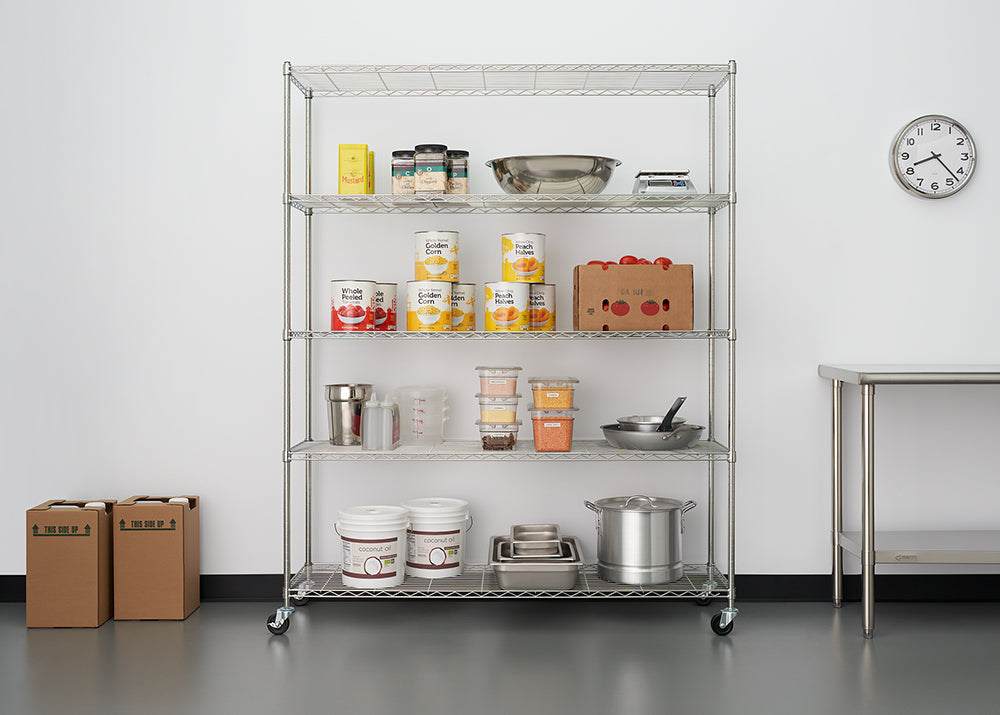 A five-shelf chrome wire shelving unit on casters is centered against a white wall and stocked with various food storage and cooking supplies, including canned goods, dry goods, mixing bowls, and pots. To the left are two cardboard boxes, and a stainless steel utility table is visible on the right next to a wall clock.