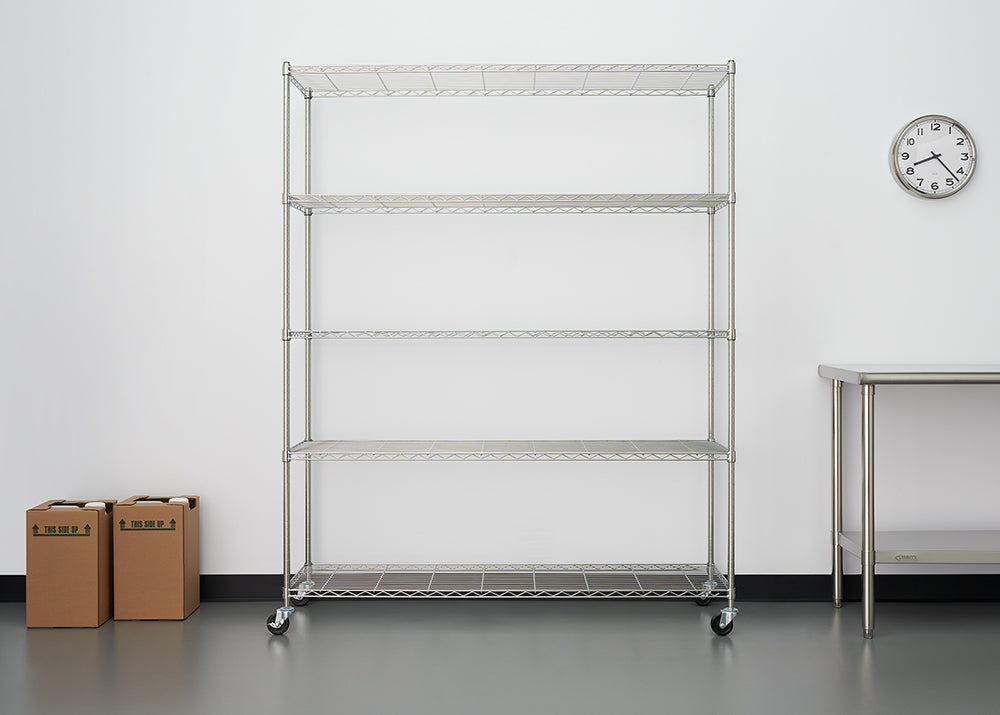 A five-shelf, rolling, wire shelving unit stands centered against a white wall in what appears to be a commercial kitchen or storage area. To the right is a stainless steel table, and two small cardboard boxes sit on the floor to the left.