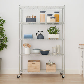 A five-shelf chrome wire shelving unit on casters is neatly organized in a bright room, holding kitchen items, pantry staples in clear containers, a plant, and wooden storage crates. To the left is a large potted faux tree, and to the right is a white kitchen cart with a wooden top.