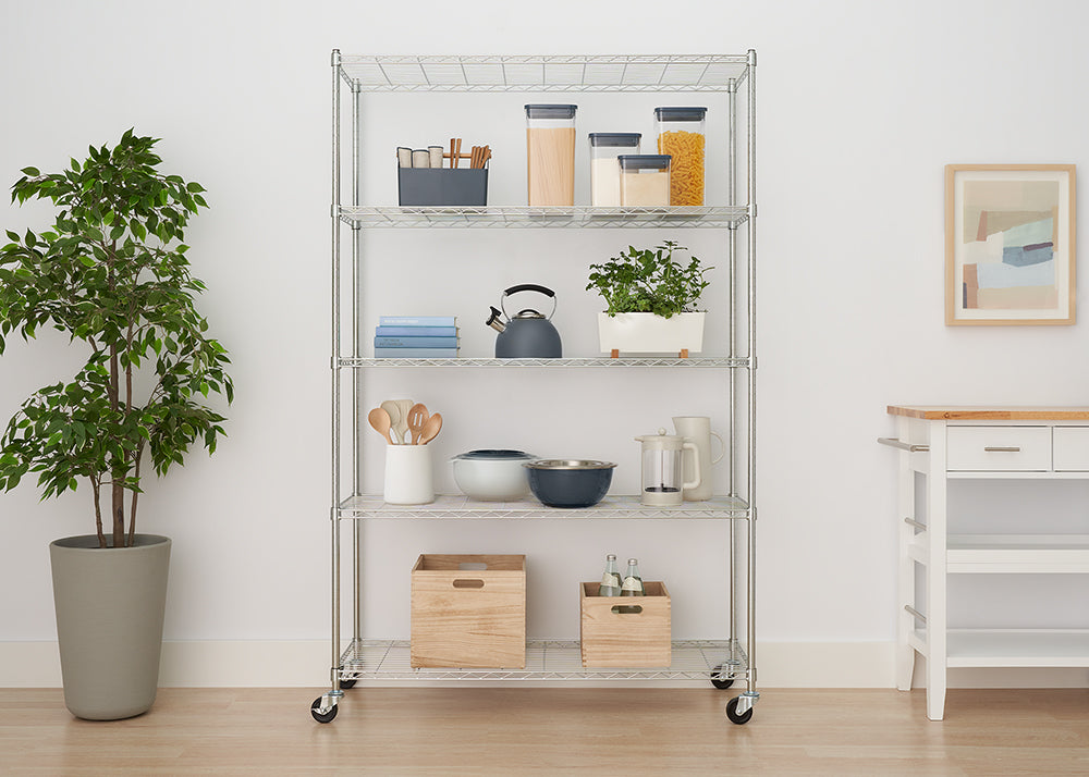 A five-shelf chrome wire shelving unit on casters is neatly organized in a bright room, holding kitchen items, pantry staples in clear containers, a plant, and wooden storage crates. To the left is a large potted faux tree, and to the right is a white kitchen cart with a wooden top.
