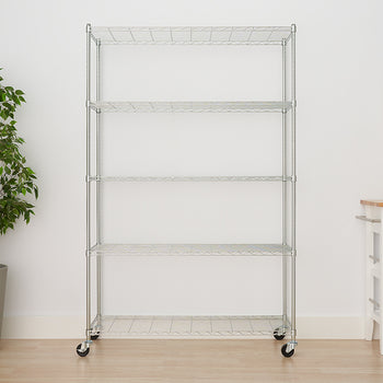 A large, five-tier chrome wire shelving unit with four locking casters stands centered against a white wall. To the left is a tall potted faux plant, and to the right is a small white and wood accent table with a framed abstract print hanging above it.