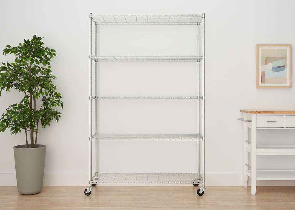 A large, five-tier chrome wire shelving unit with four locking casters stands centered against a white wall. To the left is a tall potted faux plant, and to the right is a small white and wood accent table with a framed abstract print hanging above it.