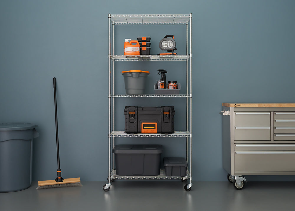 A tall, chrome wire shelving unit on wheels is centered against a blue-gray wall in a garage or workshop. It holds various black and orange storage containers, cleaning supplies, a work light, and buckets, organizing tools and supplies next to a large metal rolling workbench.