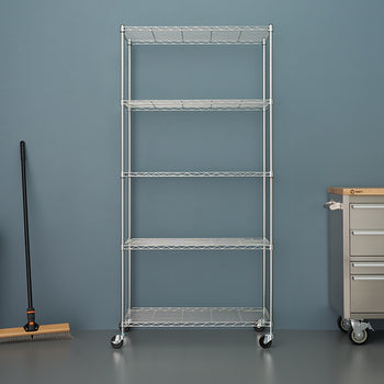 A five-tier, chrome wire shelving unit on wheels stands centered in a garage or workshop setting. To the left is a gray trash bin and a push broom, and to the right is a rolling stainless steel workbench or tool chest with a wooden top.