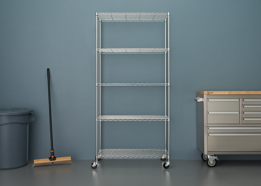A five-tier, chrome wire shelving unit on wheels stands centered in a garage or workshop setting. To the left is a gray trash bin and a push broom, and to the right is a rolling stainless steel workbench or tool chest with a wooden top.