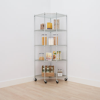 A tall, five-tier chrome wire corner shelving unit with wheels is positioned in the corner of a white-walled room with a light wood floor. The shelves are neatly stocked with various pantry items, including clear containers of pasta and flour, boxes of cereals and dry goods, and bottles of drinks on the bottom shelf.