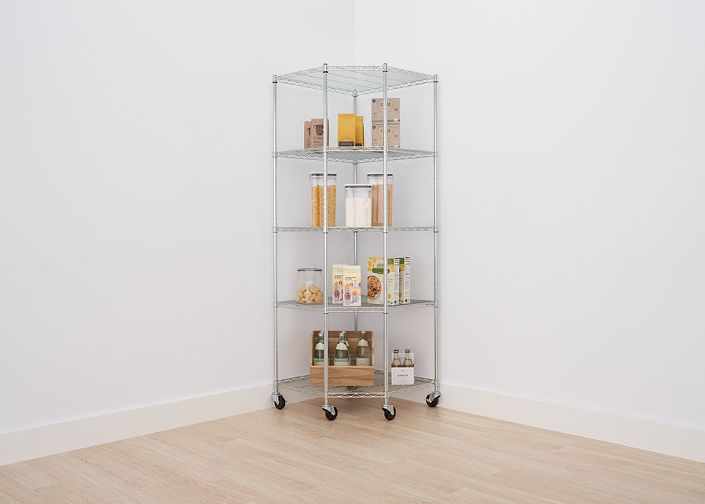 A tall, five-tier chrome wire corner shelving unit with wheels is positioned in the corner of a white-walled room with a light wood floor. The shelves are neatly stocked with various pantry items, including clear containers of pasta and flour, boxes of cereals and dry goods, and bottles of drinks on the bottom shelf.