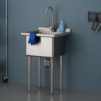 A freestanding, stainless steel utility sink with four metal legs is centered against a dark blue wall. The sink has a curved faucet, a bottle of hand soap, and a pair of blue rubber gloves draped over the front, suggesting use in a garage, laundry room, or workshop.