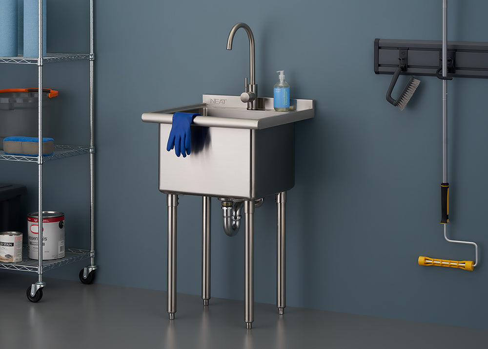 A freestanding, stainless steel utility sink with four metal legs is centered against a dark blue wall. The sink has a curved faucet, a bottle of hand soap, and a pair of blue rubber gloves draped over the front, suggesting use in a garage, laundry room, or workshop.