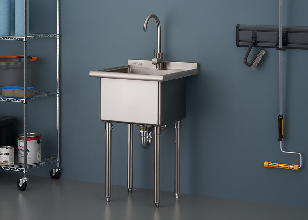 A commercial-style stainless steel utility sink with a gooseneck faucet stands on four metal legs in a garage or workshop setting. The sink is positioned against a blue-gray wall with a storage shelf unit visible on the left and cleaning tools hanging on the right.