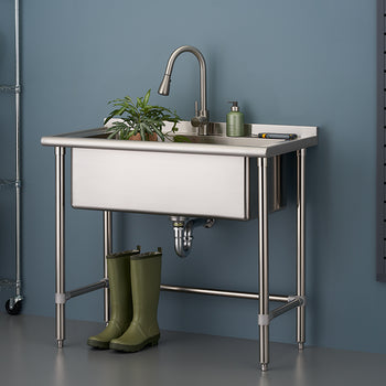 A stainless steel utility sink with a gooseneck faucet, set on four metal legs, in a laundry or garage area. A small potted plant and green soap dispenser sit on the right side of the sink, while a pair of tall green rubber boots are positioned underneath.