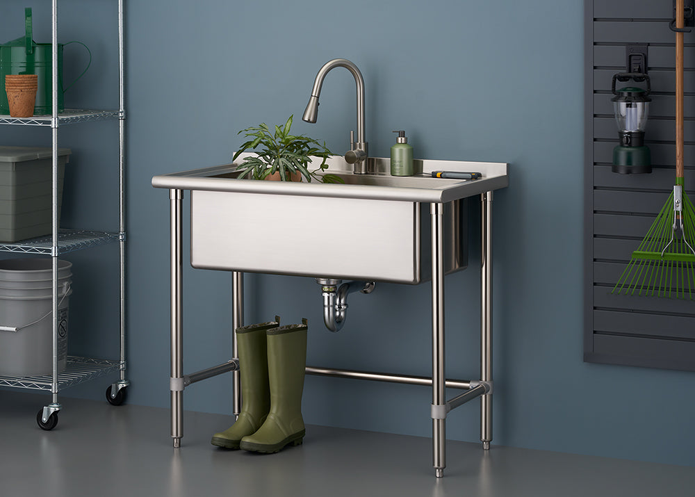 A stainless steel utility sink with a gooseneck faucet, set on four metal legs, in a laundry or garage area. A small potted plant and green soap dispenser sit on the right side of the sink, while a pair of tall green rubber boots are positioned underneath.