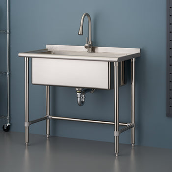 A close-up of a freestanding, single-basin stainless steel utility sink with a high-arc faucet, set up against a blue-gray wall in what appears to be a garage or laundry area. The sink has tubular stainless steel legs connected by a lower crossbar, and to the left is a wire shelving unit.