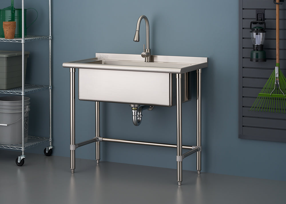 A close-up of a freestanding, single-basin stainless steel utility sink with a high-arc faucet, set up against a blue-gray wall in what appears to be a garage or laundry area. The sink has tubular stainless steel legs connected by a lower crossbar, and to the left is a wire shelving unit.