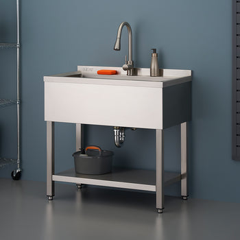 A shiny stainless steel utility sink with a tall faucet stands on metal legs, featuring a lower shelf and a small bucket placed underneath. The sink is set against a blue wall in what appears to be a garage or workshop, with wire shelving and various cleaning tools visible on the right.
