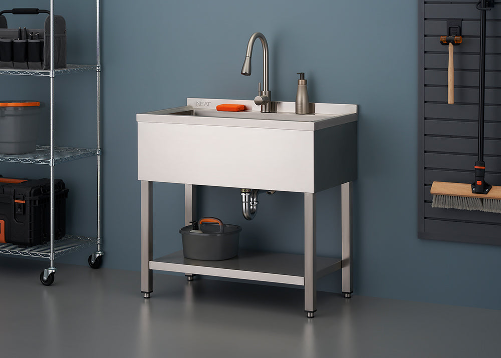 A shiny stainless steel utility sink with a tall faucet stands on metal legs, featuring a lower shelf and a small bucket placed underneath. The sink is set against a blue wall in what appears to be a garage or workshop, with wire shelving and various cleaning tools visible on the right.