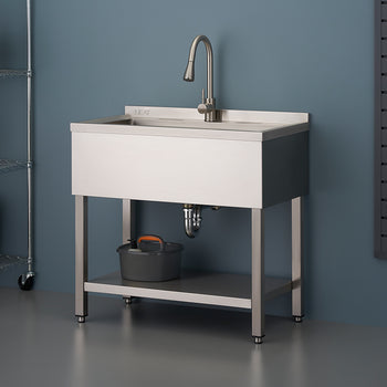 A stainless steel utility sink with a gooseneck faucet stands against a blue-gray wall in a garage or workshop setting. The sink unit features a lower shelf, a visible P-trap, and is flanked by wire shelving on the left and a wall-mounted tool panel on the right.