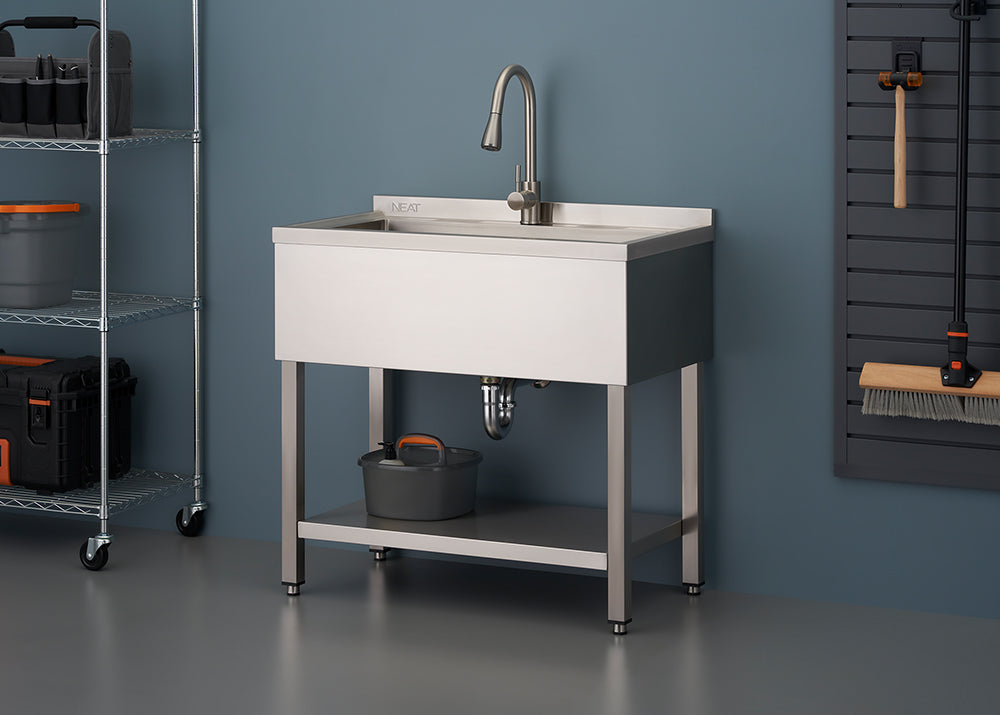A stainless steel utility sink with a gooseneck faucet stands against a blue-gray wall in a garage or workshop setting. The sink unit features a lower shelf, a visible P-trap, and is flanked by wire shelving on the left and a wall-mounted tool panel on the right.