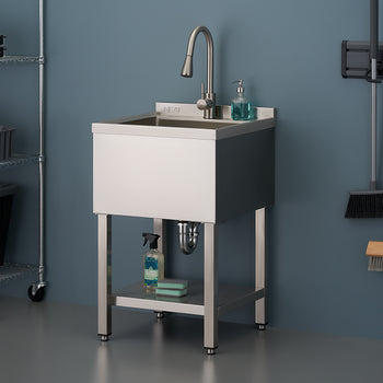 A stainless steel utility sink with a faucet and integrated backsplash stands on matching metal legs in a garage or utility room. A small shelf beneath the basin holds cleaning supplies, and metal shelving and hanging cleaning tools are visible in the background.