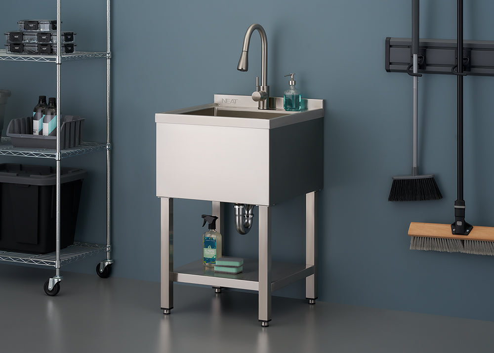 A stainless steel utility sink with a faucet and integrated backsplash stands on matching metal legs in a garage or utility room. A small shelf beneath the basin holds cleaning supplies, and metal shelving and hanging cleaning tools are visible in the background.