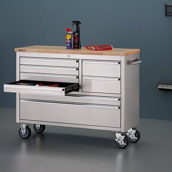 A stainless steel mobile tool workbench with a light wood top is shown in a garage setting with all-gray walls and flooring. The bottom-left shallow drawer is pulled open, displaying organized hand tools inside. On top of the wooden surface sit spray cans and a red tool organizer tray, while wall-mounted cleaning tools hang nearby on the right.