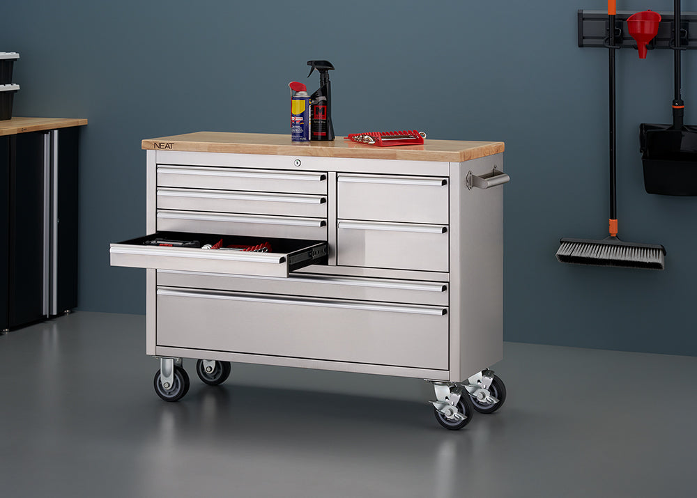 A stainless steel mobile tool workbench with a light wood top is shown in a garage setting with all-gray walls and flooring. The bottom-left shallow drawer is pulled open, displaying organized hand tools inside. On top of the wooden surface sit spray cans and a red tool organizer tray, while wall-mounted cleaning tools hang nearby on the right.