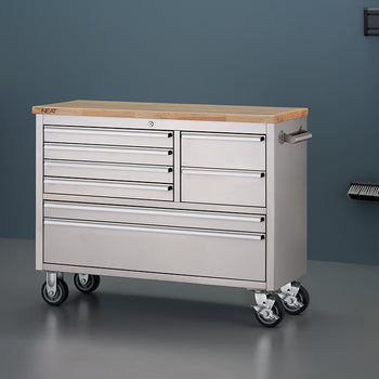 A stainless steel mobile tool workbench with a light wood top is shown in a garage setting with all-gray walls and flooring. The unit features all-closed drawers, four heavy-duty caster wheels, and a side pull handle, with a clean surface free of any items.