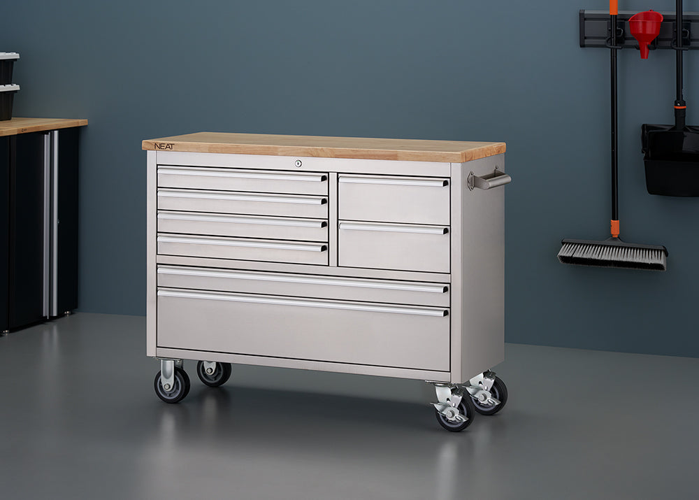 A stainless steel mobile tool workbench with a light wood top is shown in a garage setting with all-gray walls and flooring. The unit features all-closed drawers, four heavy-duty caster wheels, and a side pull handle, with a clean surface free of any items.
