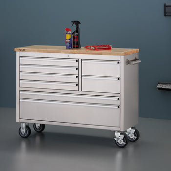 A stainless steel mobile tool workbench with a light wood top is shown in a workshop setting with all-gray walls and flooring. All drawers are closed, with spray bottles and a red wrench organizer resting on top, and wall-mounted cleaning tools hanging on the right wall.
