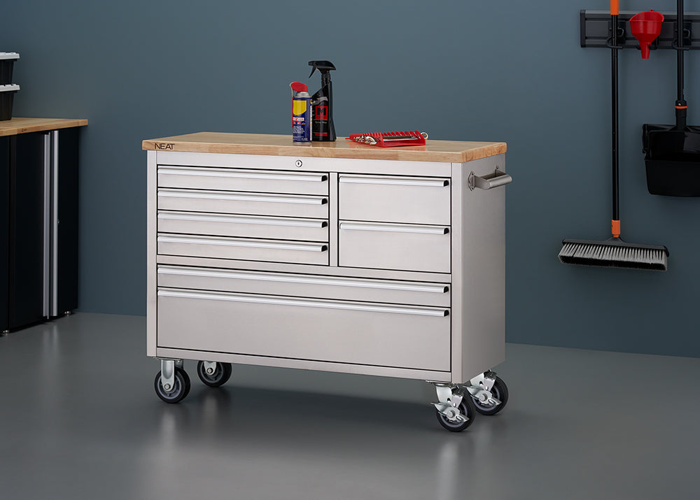 A stainless steel mobile tool workbench with a light wood top is shown in a workshop setting with all-gray walls and flooring. All drawers are closed, with spray bottles and a red wrench organizer resting on top, and wall-mounted cleaning tools hanging on the right wall.