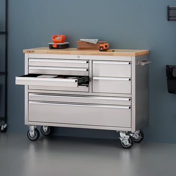 A stainless steel mobile tool workbench with a light wooden top stands against a muted blue workshop wall, mounted on heavy-duty casters. One of its upper drawers is pulled open, revealing internal storage, while a tape measure, small hardware bins, and stacked wood scraps rest on top. Surrounding the workbench are metal shelving racks on the left and wall-mounted cleaning tools on the right.