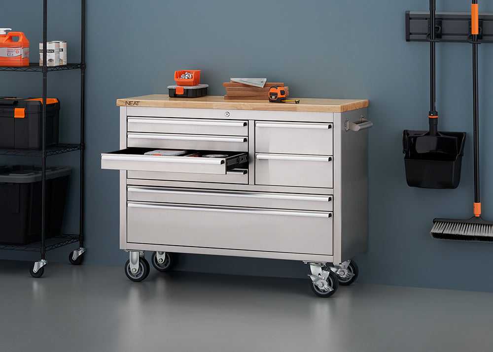 A stainless steel mobile tool workbench with a light wooden top stands against a muted blue workshop wall, mounted on heavy-duty casters. One of its upper drawers is pulled open, revealing internal storage, while a tape measure, small hardware bins, and stacked wood scraps rest on top. Surrounding the workbench are metal shelving racks on the left and wall-mounted cleaning tools on the right.